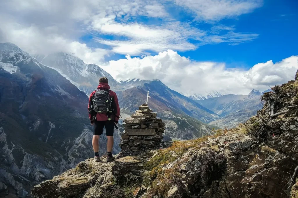 Homem com mochila e roupas de caminhada em pé sobre um cume rochoso, observando um vale alpino profundo e montanhas nevadas ao longe.