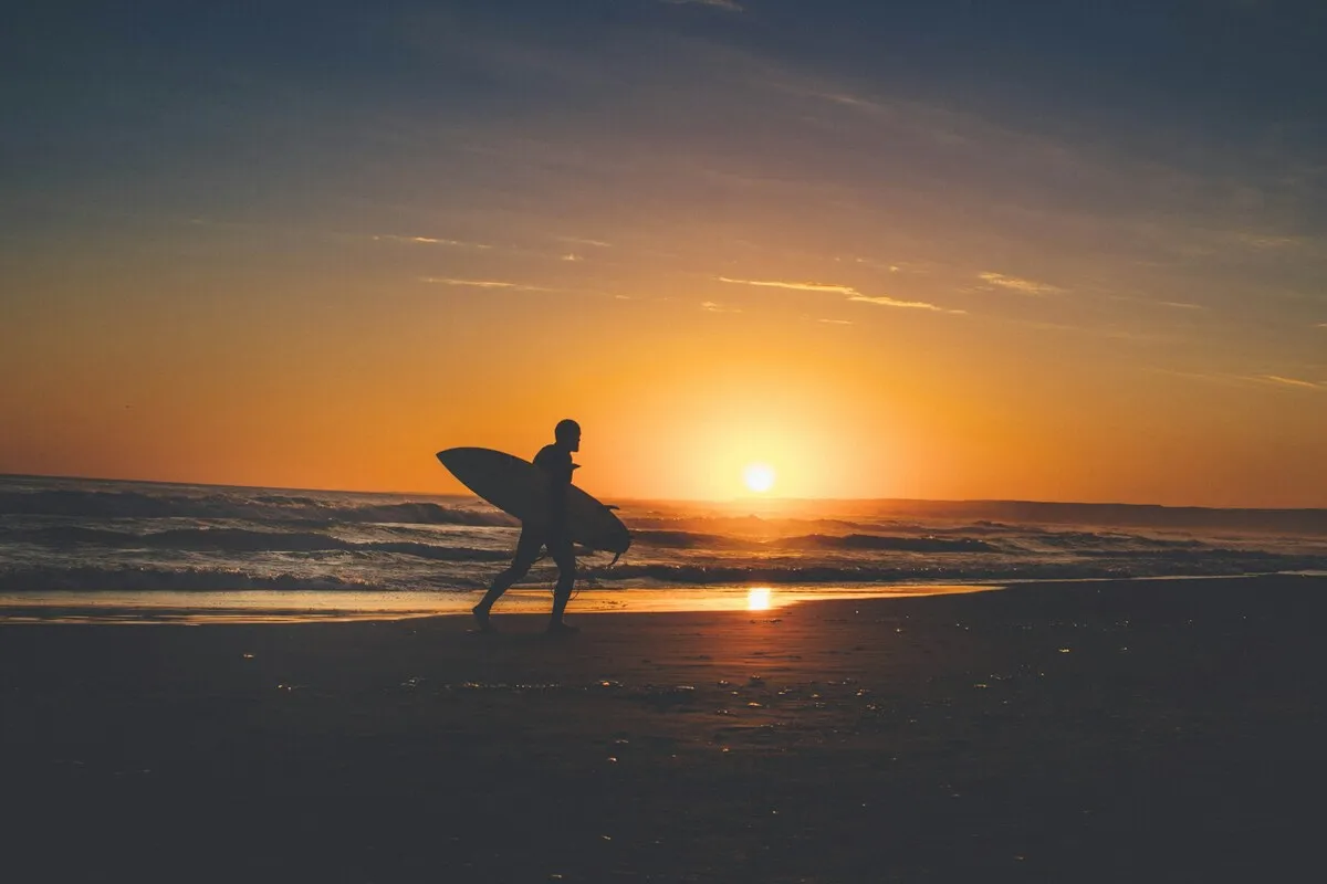 A silhueta de um surfista caminhando na praia ao pôr do sol em Cabo Polonio, Uruguai, carregando sua prancha debaixo do braço.