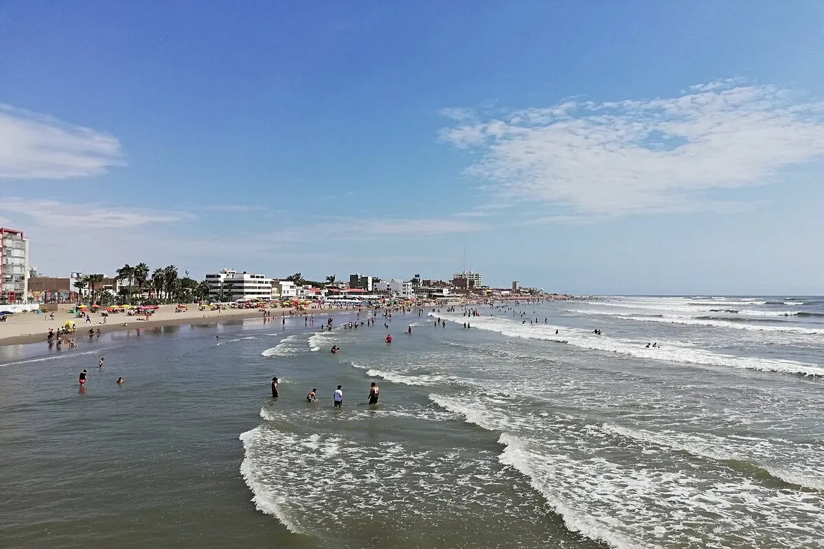 Vista da Praia de Pimentel em Chiclayo, Peru, com banhistas aproveitando o mar e construções urbanas à beira-mar.