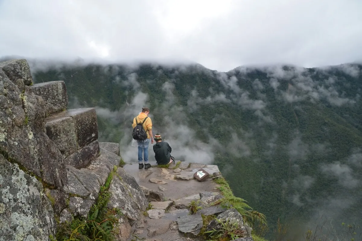 Duas pessoas em um penhasco rochoso na montanha Huayna Picchu, observando um vale montanhoso coberto por névoa e nuvens baixas.