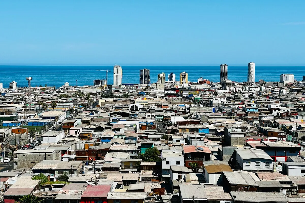 Vista panorâmica da cidade de Iquique, Chile, mostrando um bairro com casas de baixa renda em primeiro plano e uma orla com arranha-céus modernos à beira do oceano Pacífico.