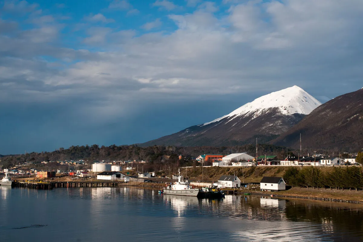 Vista de nível do solo da orla marítima de Puerto Williams, Chile. Barcos atracados perto de cais de madeira com edifícios da cidade ao fundo, dominados por um pico nevado sob um céu azul e dramático.