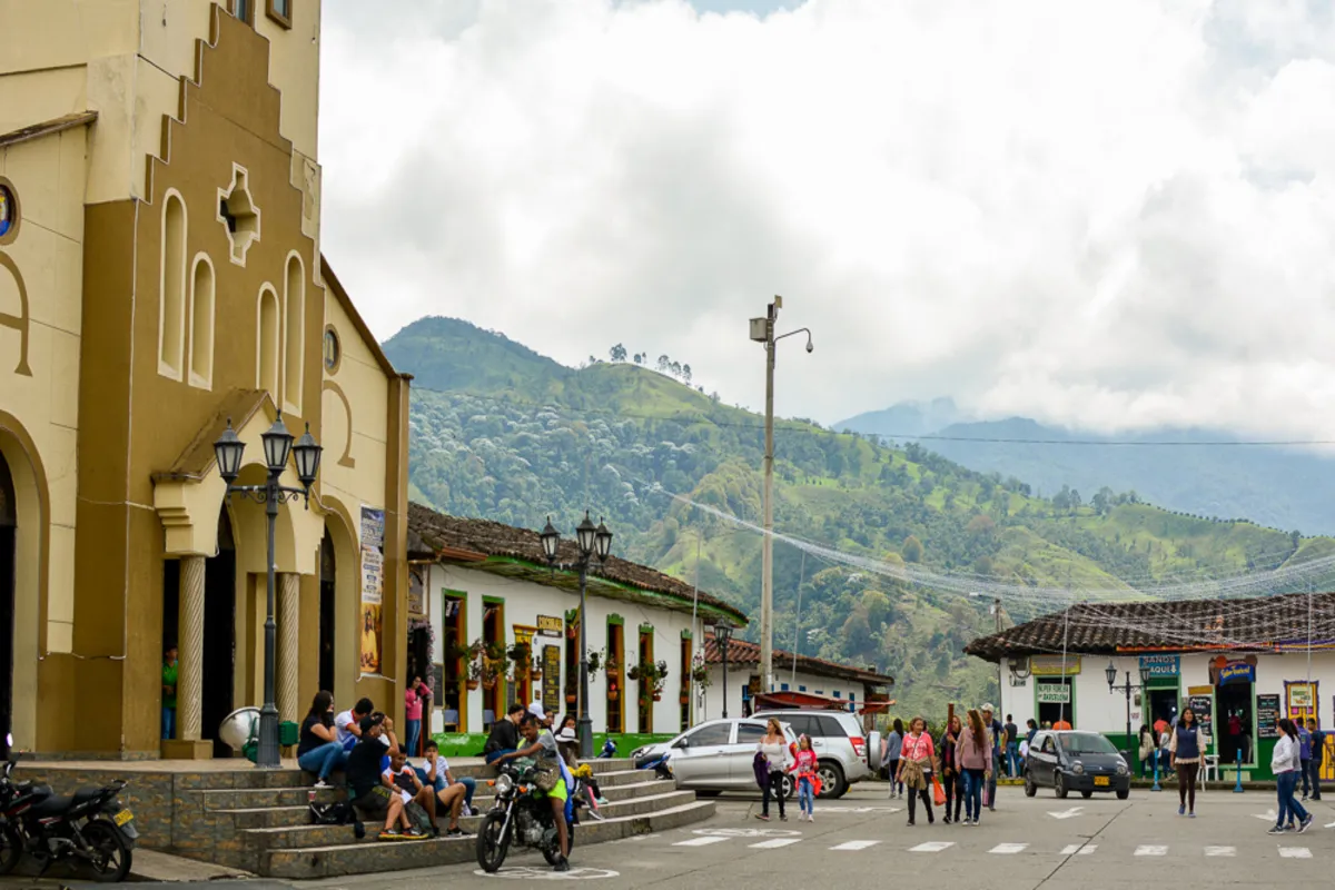 Uma vista da praça principal de Salento, Colômbia, com a igreja de arquitetura colonial, montanhas verdes ao fundo e pessoas caminhando.