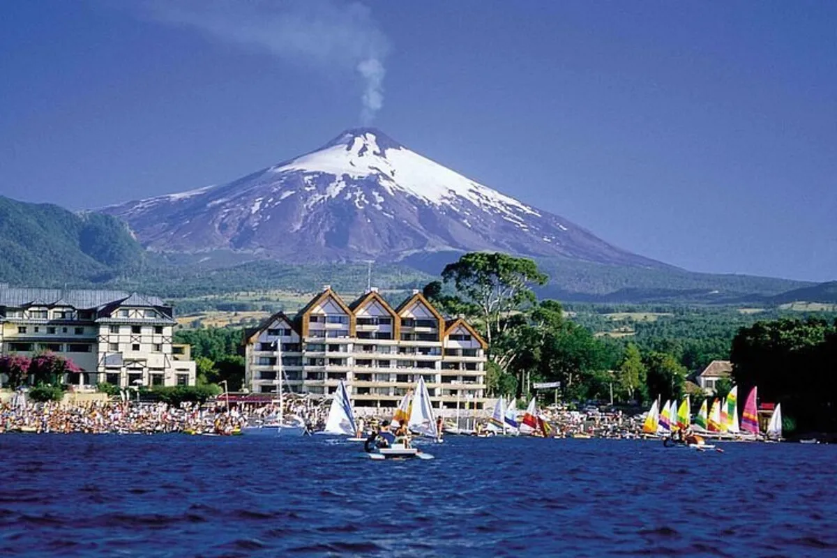 O vulcão Villarrica com seu cume coberto de neve e fumaça, ao fundo, com o lago Villarrica em primeiro plano, onde veleiros e barcos navegam em um dia ensolarado.
