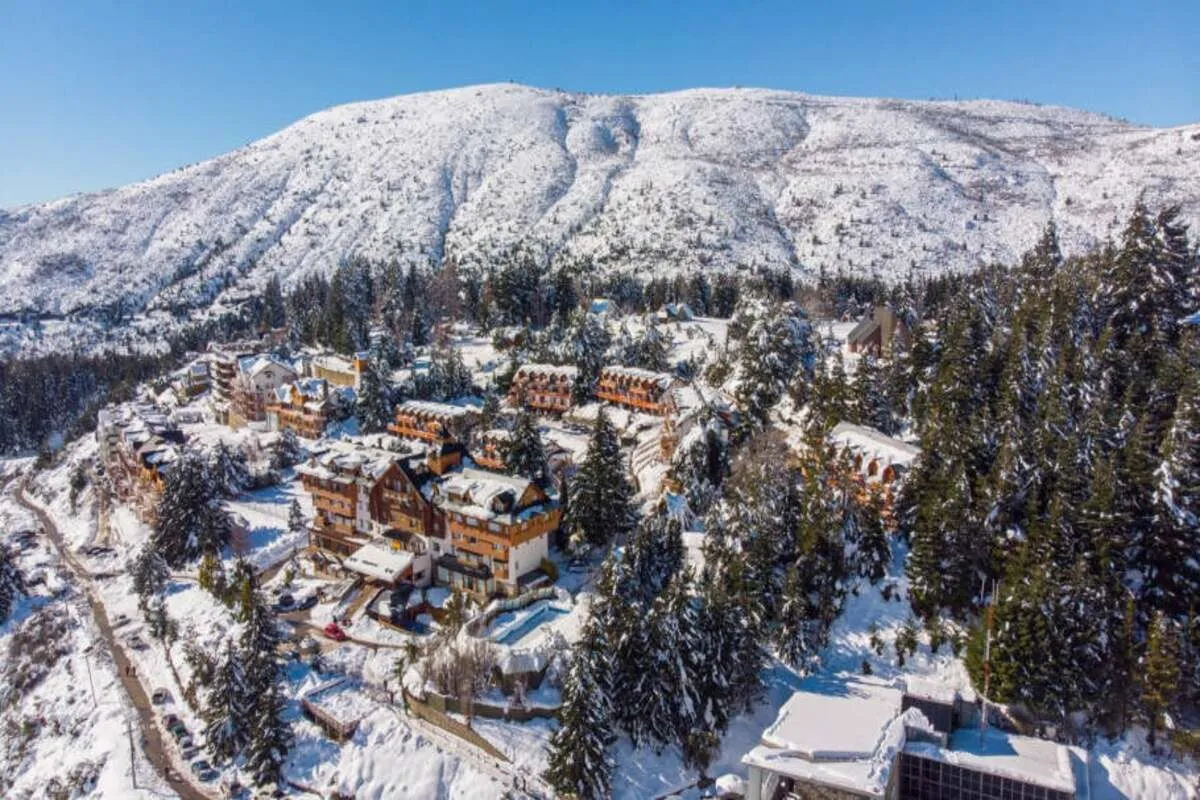 Vista aérea de um complexo de hotéis e chalés em Bariloche, cercado por pinheiros e montanhas cobertas de neve, com um céu azul e limpo.
