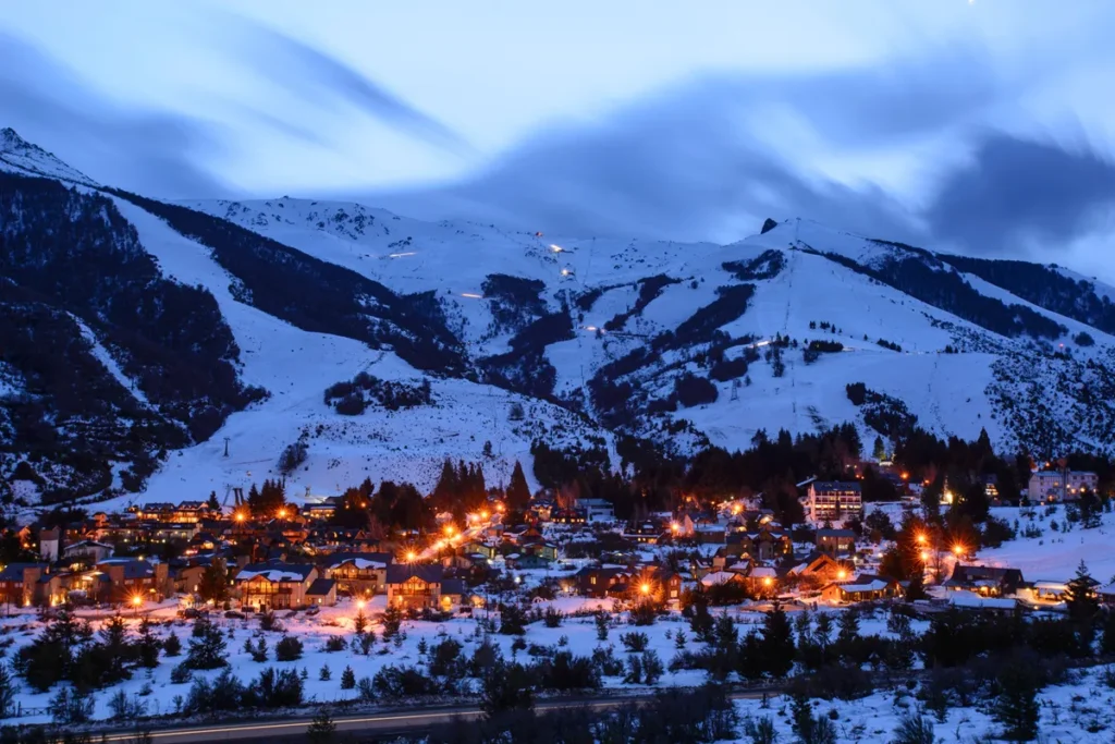 Vista panorâmica de Bariloche ao entardecer, com luzes douradas iluminando a cidade coberta de neve e montanhas brancas ao fundo na Patagônia argentina.