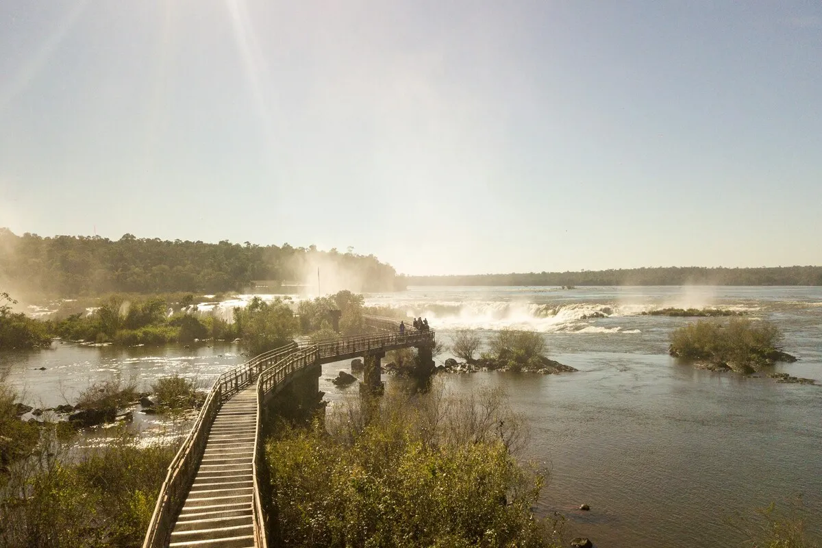 Passarela de madeira curva sobre o Rio Iguaçu, com turistas, levando a vistas das quedas d'água e névoa, sob sol forte no Parque Nacional Iguazú, Argentina.
