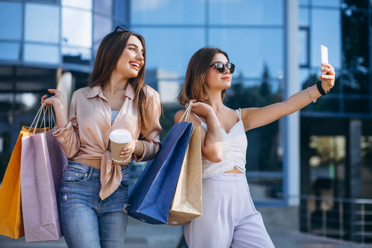 Duas amigas com sacolas de compras, uma delas tirando uma selfie, em frente a uma fachada de prédio moderno e espelhado.