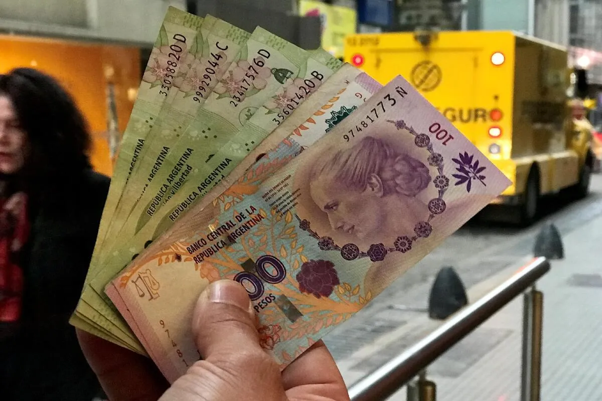 Pessoa segurando várias cédulas de pesos argentinos, incluindo notas de 100 e 200 pesos, em uma rua urbana da Argentina.