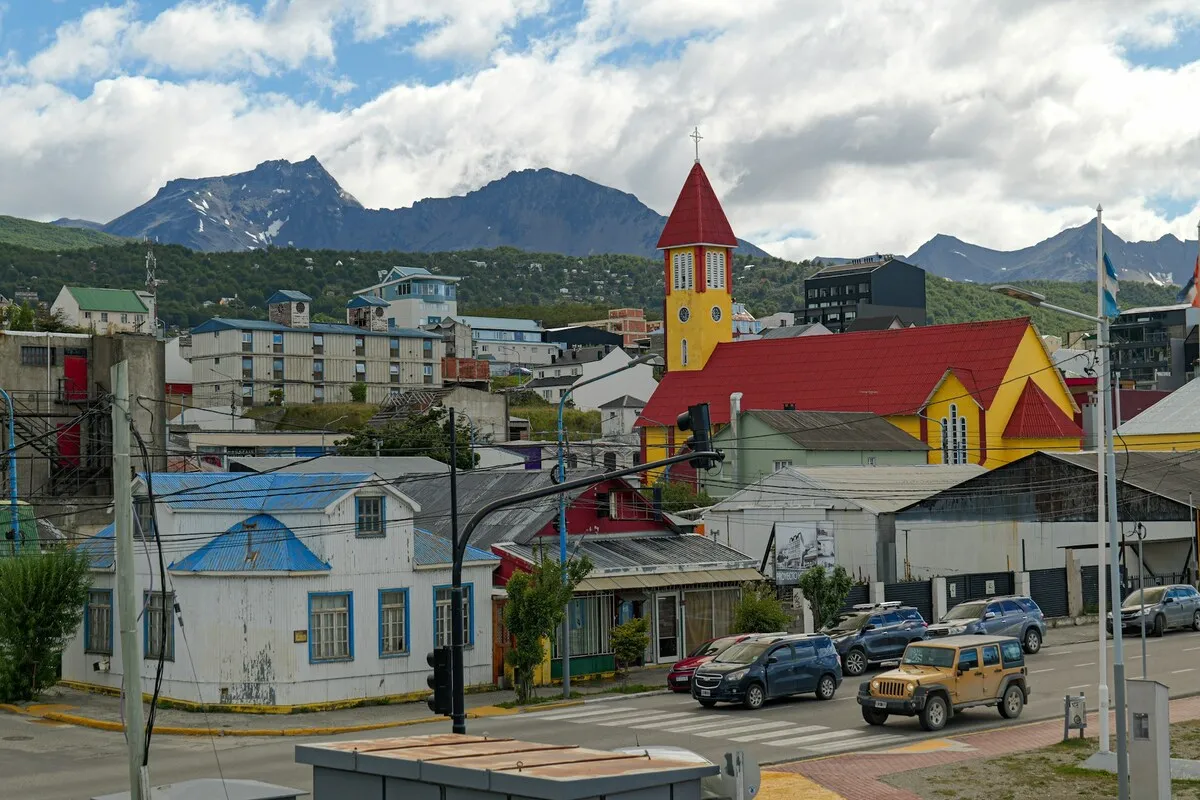A foto mostra uma vista de uma parte da cidade de Ushuaia, na Argentina. No centro da imagem, destaca-se a igreja com um telhado vermelho e a torre do sino em vermelho e amarelo. O cenário é urbano, com prédios de diferentes cores e estilos, além de carros em uma rua. Ao fundo, imponentes montanhas com picos nevados dominam a paisagem, conectando a cidade à natureza da Patagônia. O céu está parcialmente nublado, mas a luz do sol ilumina a cidade.