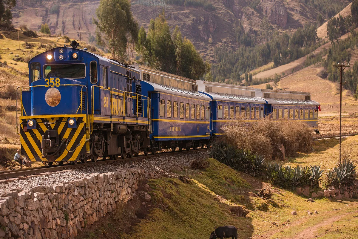 O exterior de um trem moderno com janelas grandes, de cor preta e cinza, viajando por uma paisagem montanhosa e verde no Peru. O trem está em um trilho próximo a um rio.