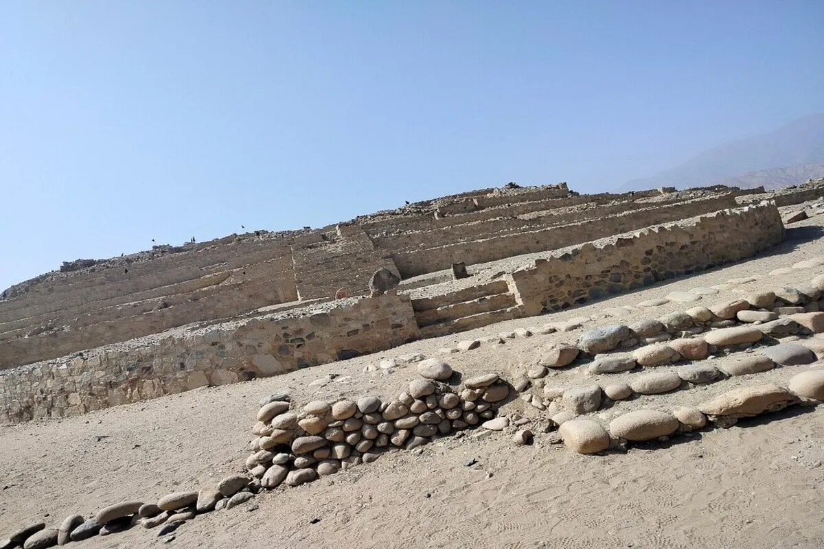 Vista lateral da base de uma pirâmide escalonada no sítio arqueológico de Caral, Peru. A estrutura é feita de pedra e terra, com vários terraços inclinados para a esquerda. Em primeiro plano, uma parede baixa de pedras redondas de rio e solo árido dominam a cena. O fundo mostra a paisagem de colinas secas sob um céu claro.