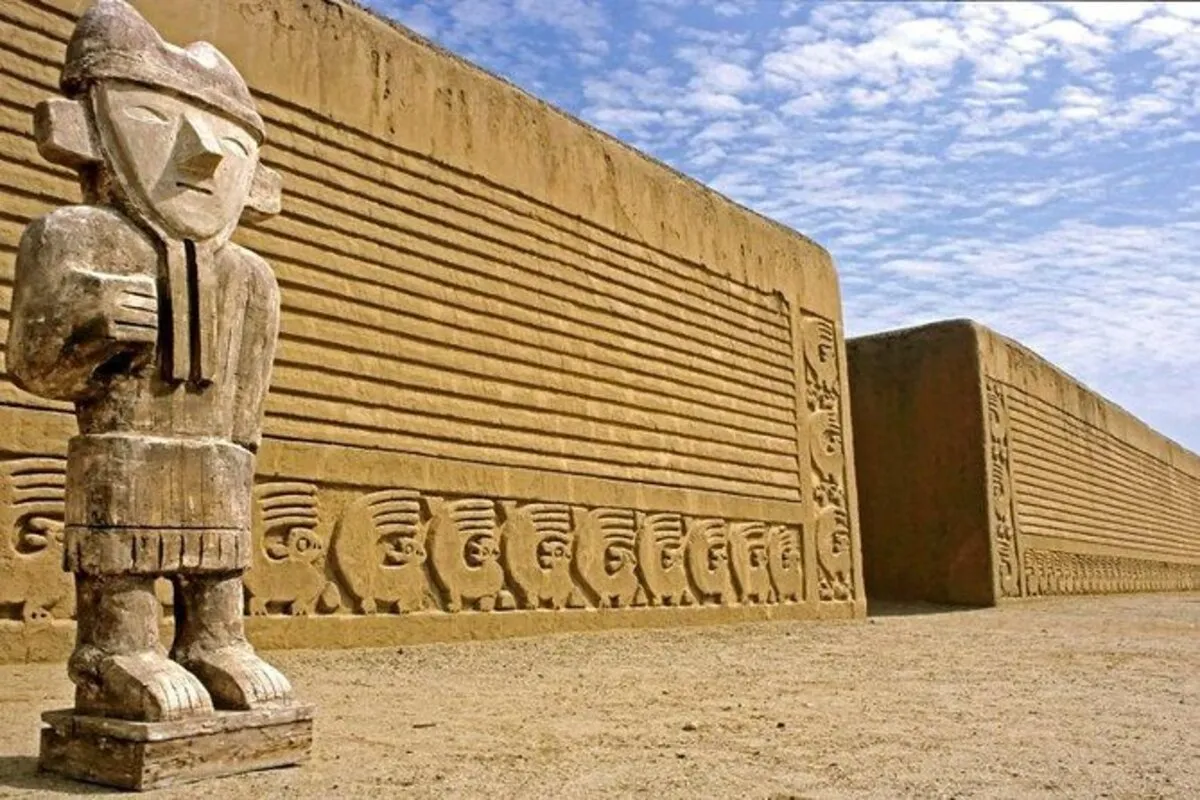 Estátua de madeira (ou adobe) de figura humana em primeiro plano, ao lado de muro de adobe com frisos em relevo em Chan Chan, Peru.