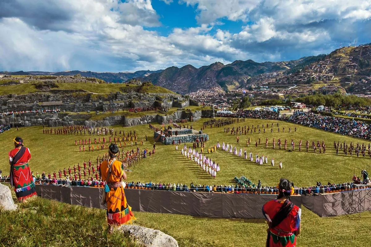 Vista panorâmica da reconstituição do Inti Raymi com centenas de figurantes em campo aberto, nas ruínas de Sacsayhuamán em Cusco, com montanhas e a cidade ao fundo.