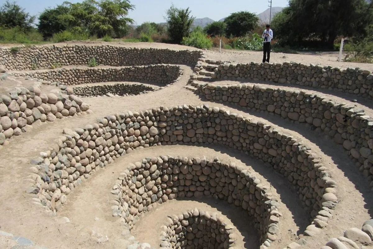 Vista em plano médio dos Aquedutos de Cantalloc (Puquios) da civilização Nazca, no Peru. São estruturas subterrâneas em espiral, com paredes construídas com pedras de rio empilhadas e secas. A rampa em formato de caracol desce em círculos no solo árido. Um homem de camisa branca e calças escuras está em pé no topo de uma das estruturas. O ambiente é seco com alguma vegetação ao fundo.