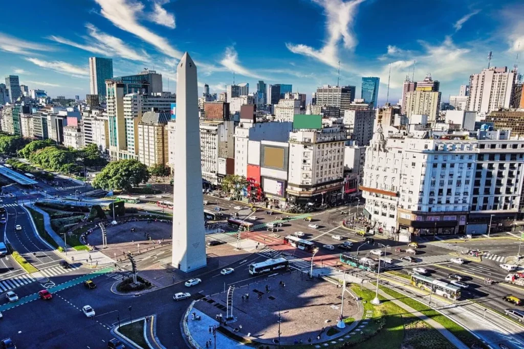 Vista aérea em dia de sol do Obelisco de Buenos Aires, um monumento alto e branco, no centro de uma movimentada intersecção de avenidas largas. O Obelisco é cercado por arranha-céus e edifícios clássicos sob um céu azul vibrante.