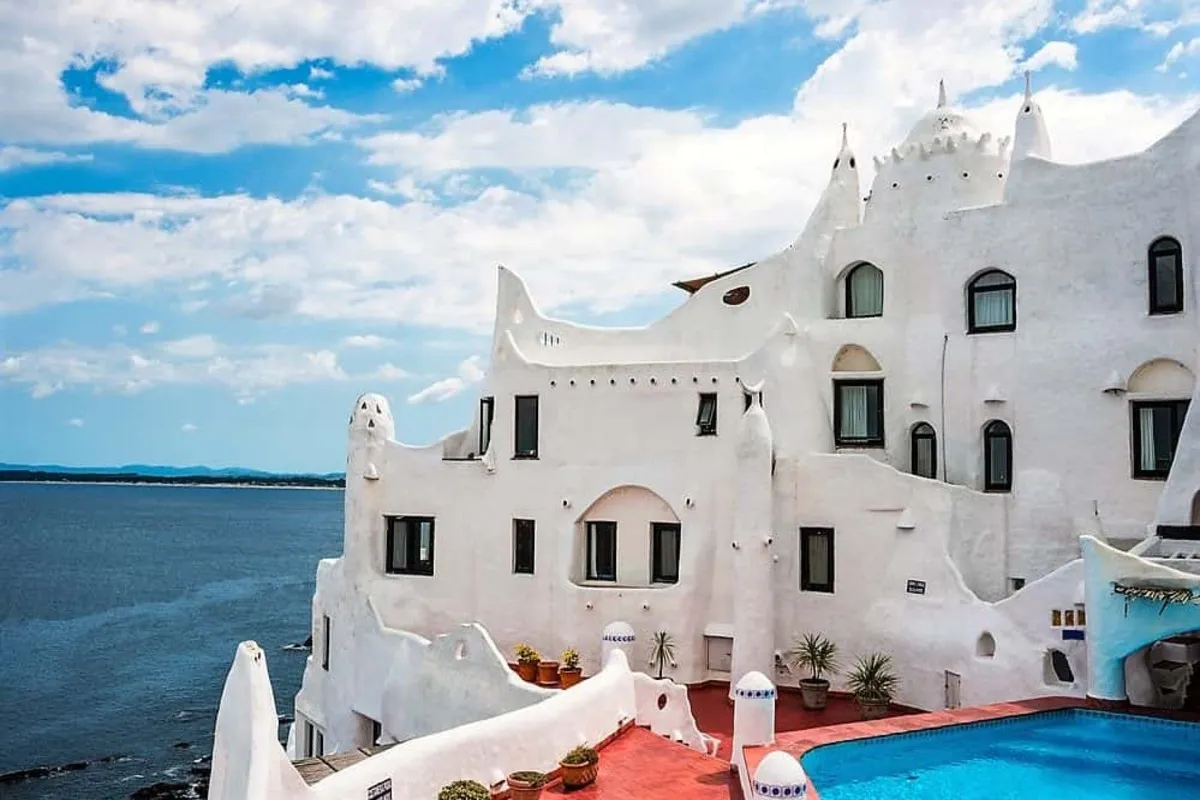 Vista frontal de Casapueblo, a arquitetura de arte branca de Carlos Páez Vilaró, sob um céu azul com nuvens brancas, destacando a piscina e a vista para o Oceano Atlântico.