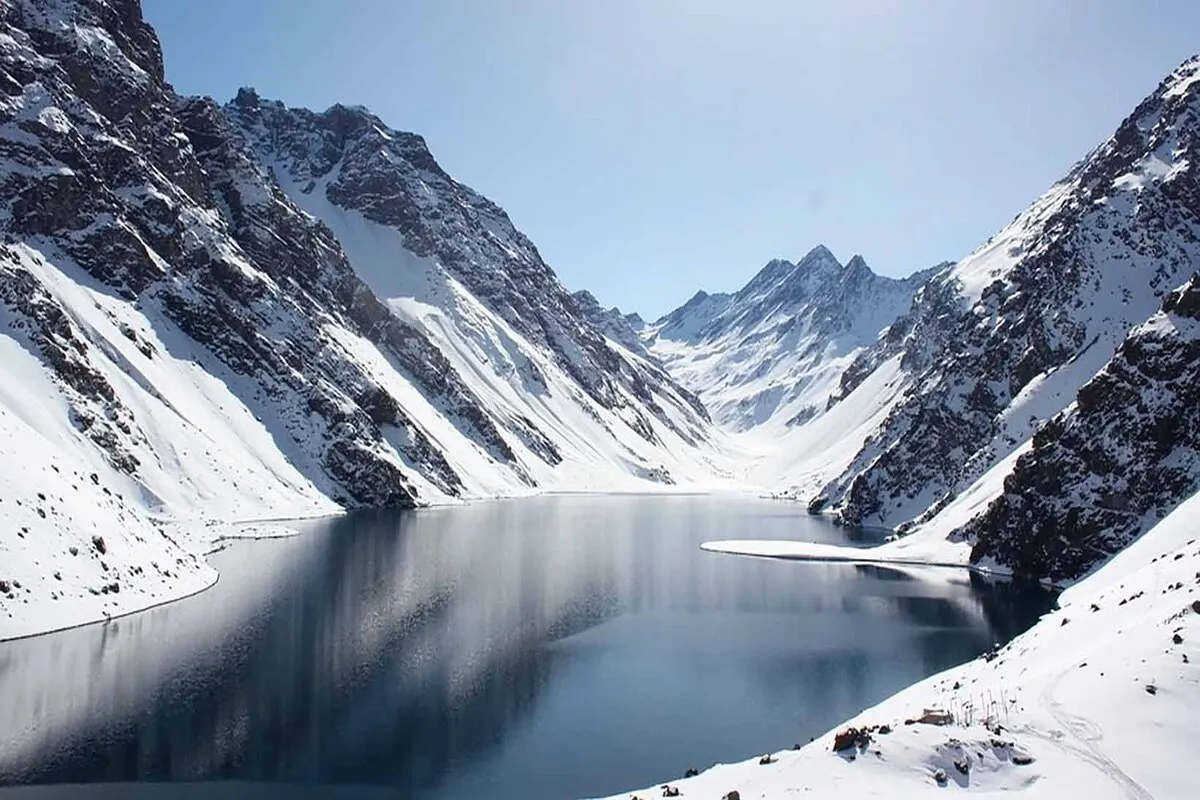 Vista panorâmica do Embalse El Yeso (ou lago de alta altitude) no Chile, cercado por montanhas brancas e nevadas. A água azul-escura e espelhada no centro reflete o céu claro de inverno, destacando a beleza da Cordilheira dos Andes sob a neve.