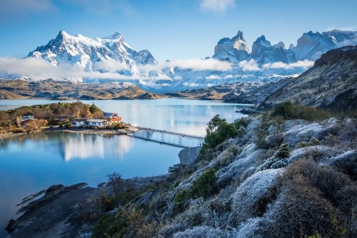 Vista panorâmica do Lago Pehoé em Torres del Paine, Chile, no inverno, com as montanhas nevadas dos Cuernos del Paine ao fundo, uma ponte e vegetação coberta de geada (neve leve) em primeiro plano.