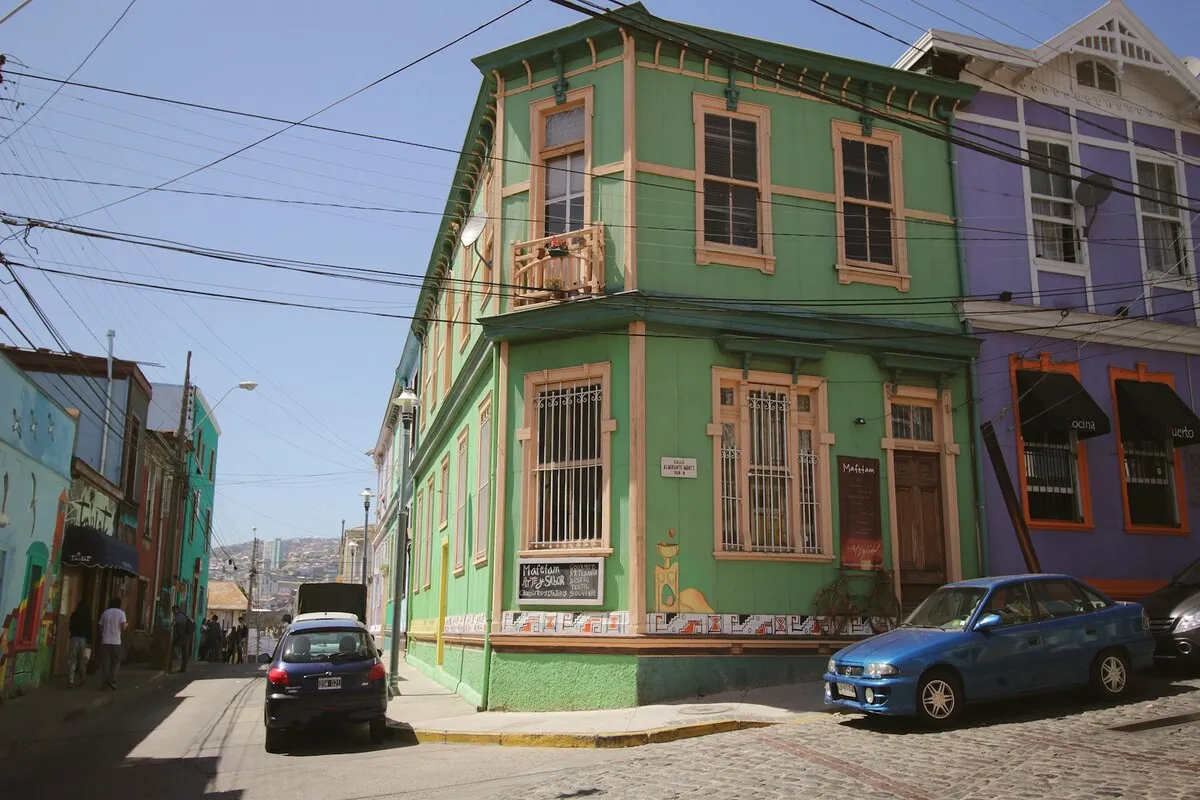 Esquina de rua de paralelepípedos em Valparaíso, Chile, destacando uma casa histórica de três andares pintada em verde-claro com detalhes em bege, e um carro azul estacionado.