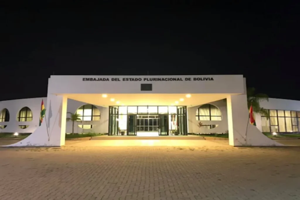Vista noturna da fachada de um edifício moderno e branco, com uma grande entrada coberta e iluminada. O letreiro no topo diz "EMBAJADA DEL ESTADO PLURINACIONAL DE BOLIVIA". Bandeiras bolivianas em pequenos mastros estão em ambos os lados da entrada.