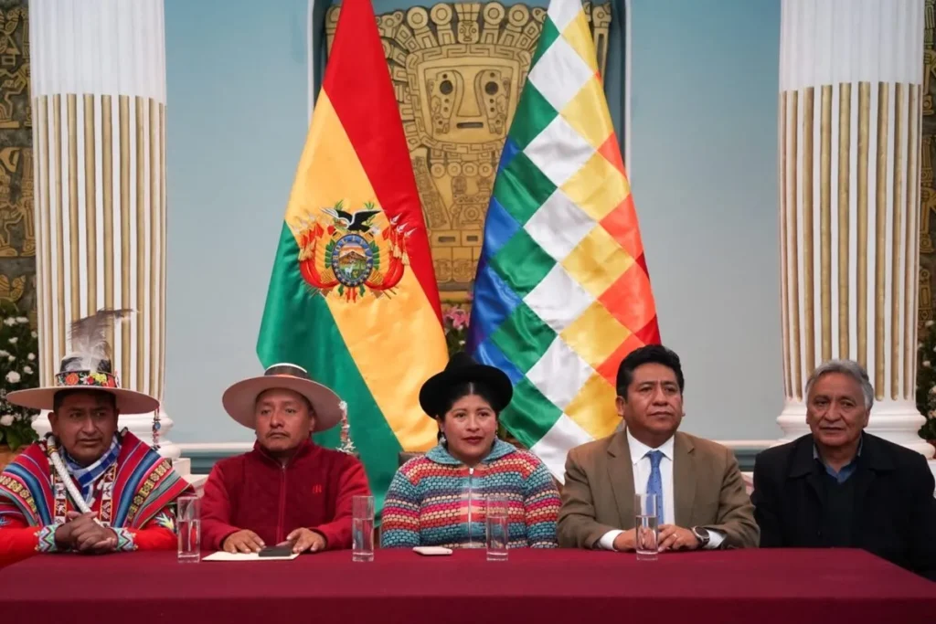 Cinco representantes, incluindo membros em trajes tradicionais indígenas e em trajes de negócios, sentados em uma mesa durante um evento formal. Ao fundo, as bandeiras nacional da Bolívia e a Wiphala estão expostas, sob um painel com um símbolo andino.