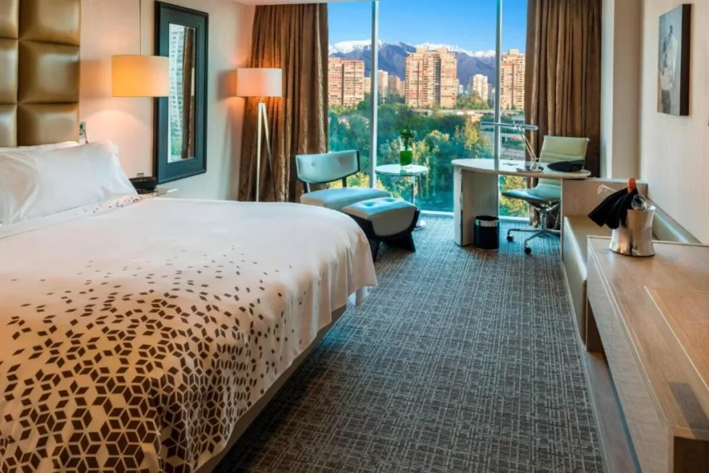 Interior de quarto de hotel de luxo em Santiago, Chile, com cama king size, decoração em tons terrosos, e uma janela panorâmica mostrando o skyline da cidade em primeiro plano e as montanhas da Cordilheira dos Andes cobertas de neve ao fundo.
