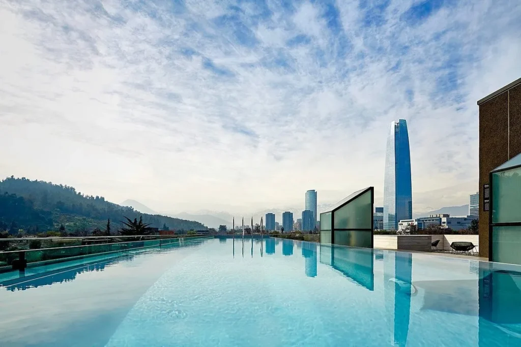 Piscina de borda infinita em um rooftop de hotel de luxo em Santiago, Chile, refletindo o céu. Ao fundo, o Skyline da cidade, incluindo a Gran Torre Santiago e as montanhas da Cordilheira dos Andes.