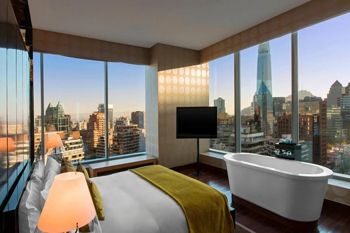 Interior de suíte de hotel de luxo em Santiago, Chile, com cama king size, banheira de imersão autônoma de frente para uma ampla janela do chão ao teto, mostrando os edifícios de Santiago, incluindo a Gran Torre Santiago.