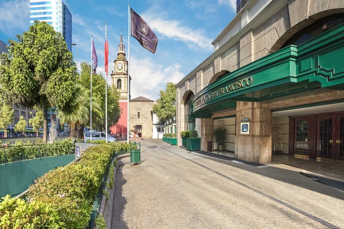 Fachada e entrada do Hotel Plaza San Francisco (ou similar) em Santiago, Chile, com um toldo verde esmeralda. Ao fundo, uma torre de igreja histórica de pedra com um relógio e a Bandeira do Chile.