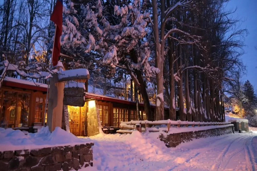 Exterior de uma pousada rústica de madeira em uma floresta no Chile durante o inverno, com o chão e árvores cobertos por neve. As janelas emitem uma luz quente que contrasta com o azul-escuro da neve e do crepúsculo.