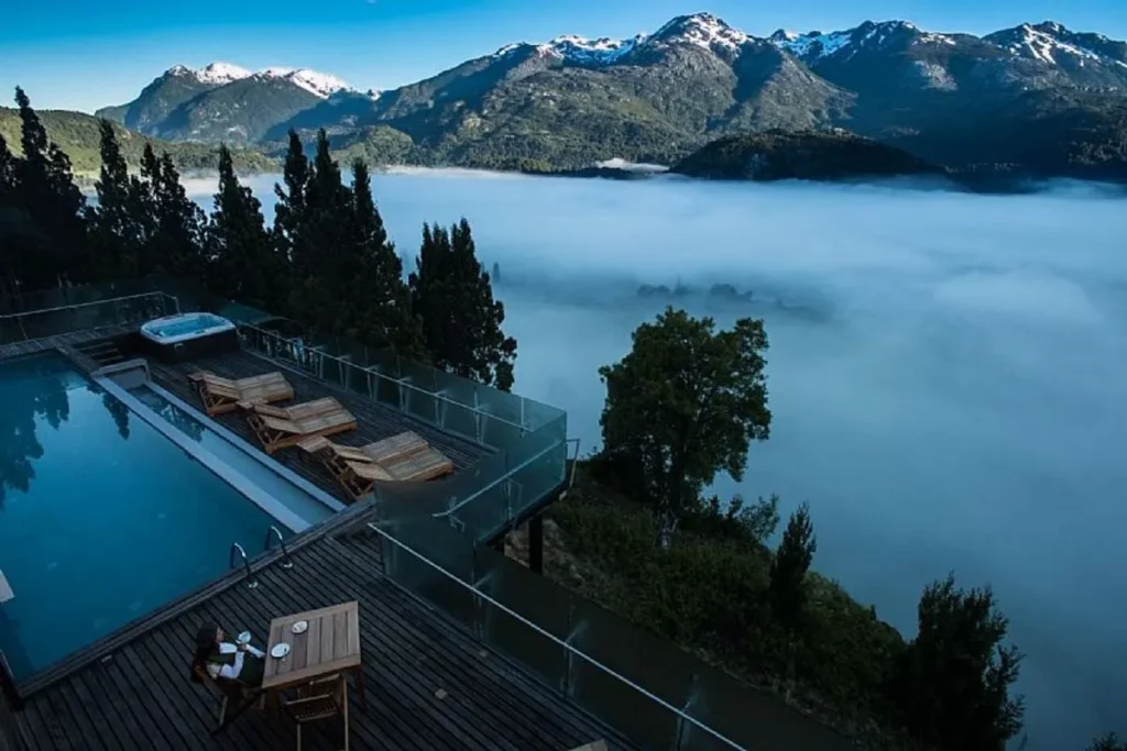 Vista aérea de uma pousada/hotel colonial no Chile com piscina e deck de madeira, com jacuzzi. O hotel está no alto de um morro, e a vista desce para um vale encoberto por um denso mar de névoa, com montanhas verdes e picos nevados ao fundo.
