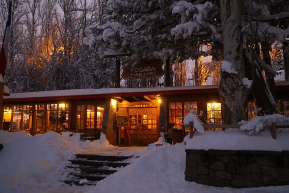 Fachada principal da Pousada Farellones rústica de madeira, coberta por neve. As janelas e a porta emitem uma luz interior quente que contrasta com o ambiente frio e azulado do inverno e o chão coberto de neve.