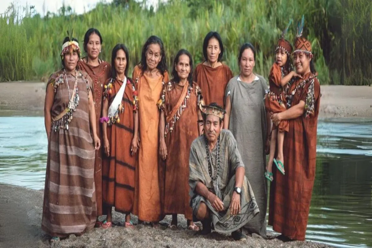 Um grupo grande de mulheres, um homem e uma criança indígena posa para uma fotografia na margem de um rio (possivelmente Amazônico), com vegetação alta ao fundo. A maioria das mulheres veste longas roupas tradicionais de algodão em tons de marrom, terracota e ocre. Um homem está agachado na frente, com um colar de miçangas, e uma mulher segura uma criança pequena. A imagem celebra a cultura e a comunidade de um povo originário.