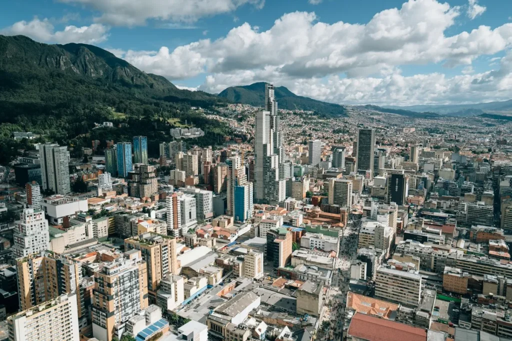 Vista aérea da densa skyline do centro de Bogotá, Colômbia, com o imponente arranha-céu BD Bacatá em destaque, cercada por morros verdes e o céu parcialmente nublado.