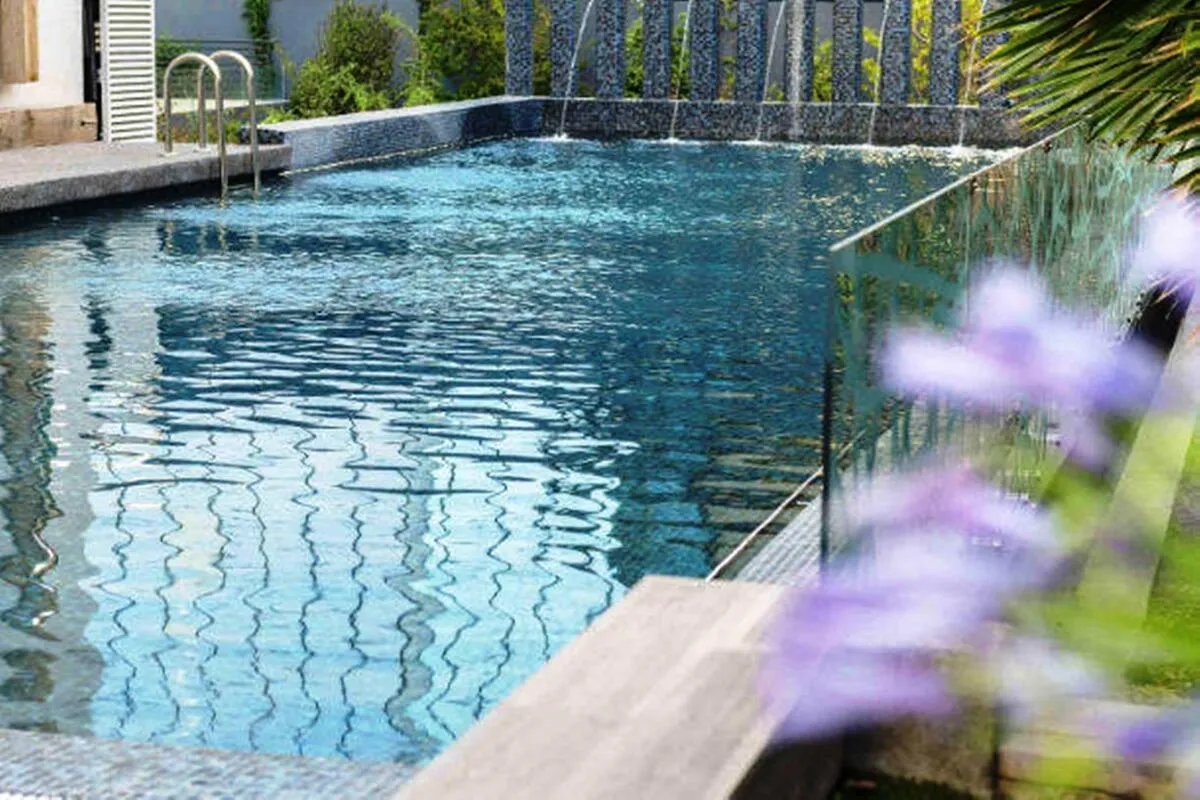 Piscina externa retangular de hotel de luxo, com água azul-escura, cercada por vidro e um muro de pedra com pequenas cascatas. O ambiente é elegante, com vegetação ao redor e design contemporâneo.