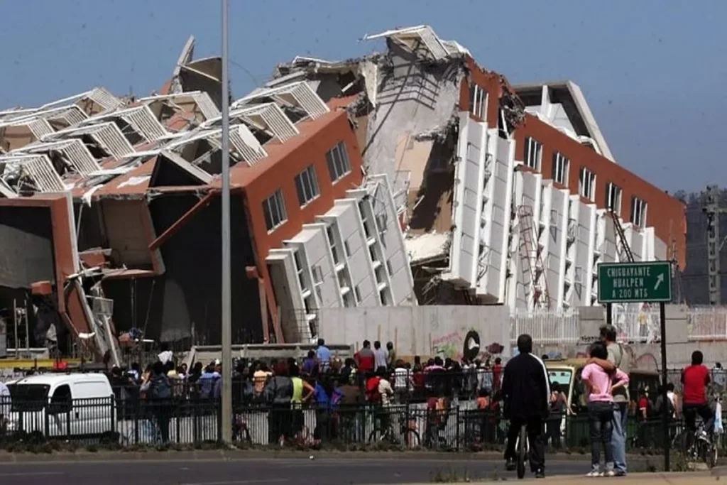 Imagem de edifício de apartamentos colapsado e danificado após um terremoto no Chile, com a estrutura inclinada. Pessoas observam a cena por trás de uma cerca na rua.