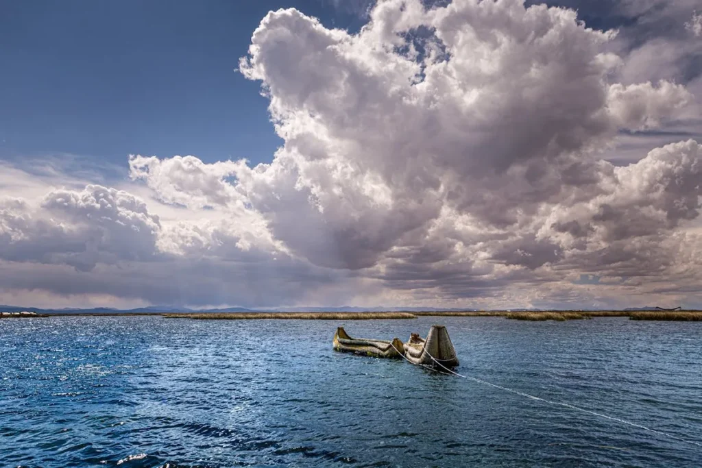 Balsa tradicional de totora (junco) flutuando no azul escuro do Lago Titicaca sob um céu dramático com grandes nuvens brancas e escuras, com um horizonte de pântanos e montanhas.