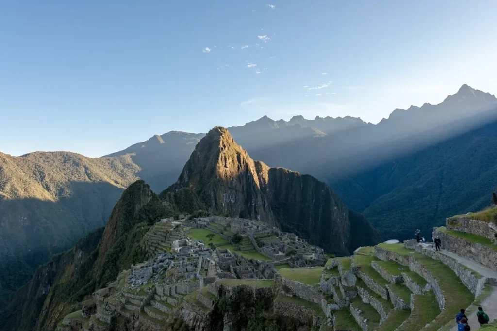 Vista panorâmica de Machu Picchu, a antiga cidadela inca, com terraços de pedra em primeiro plano, picos íngremes e raios de sol atravessando as montanhas ao fundo.