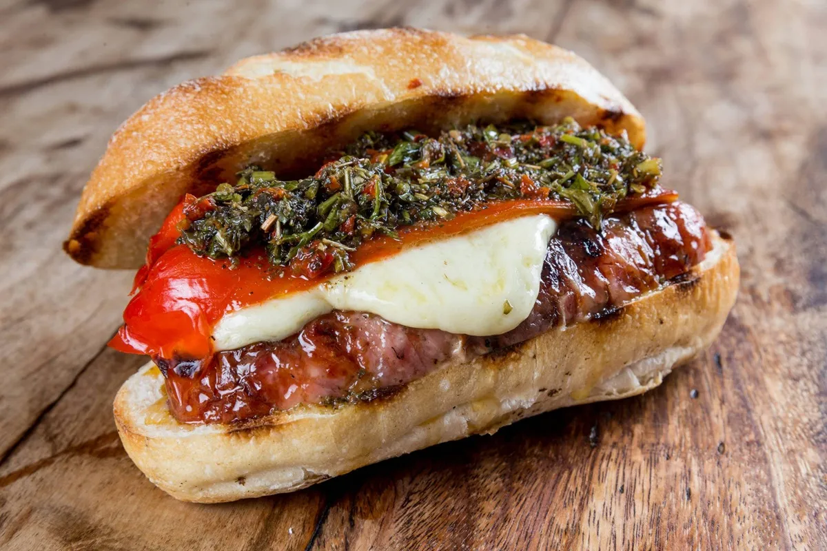 Sanduíche tradicional argentino chamado choripán, com linguiça grelhada, queijo derretido, pimentão e molho chimichurri sobre pão crocante.