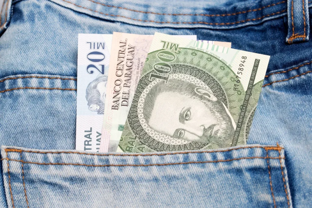 Notas de guarani paraguaio guardadas no bolso de uma calça jeans, representando câmbio e despesas de viagem.
