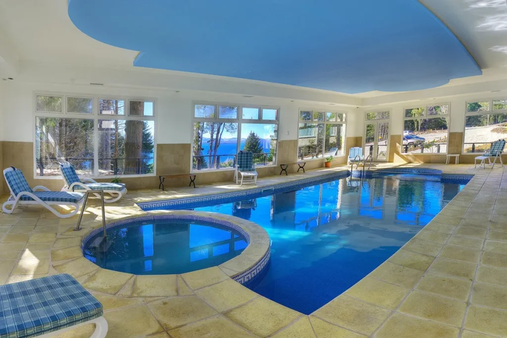 Piscina coberta de hotel em Bariloche com espreguiçadeiras e grandes janelas com vista para lago e montanhas