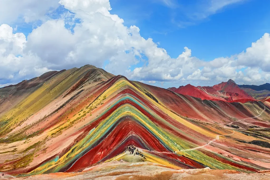 Montanha Vinicunca, no Peru, com faixas coloridas em tons de vermelho, amarelo, azul e verde sob céu azul com nuvens.