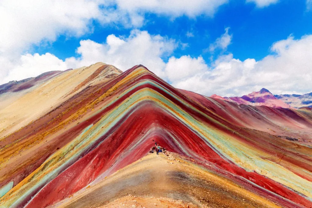 Montanha Vinicunca, no Peru, com faixas coloridas em tons de vermelho, amarelo, verde e azul sob um céu azul com nuvens.