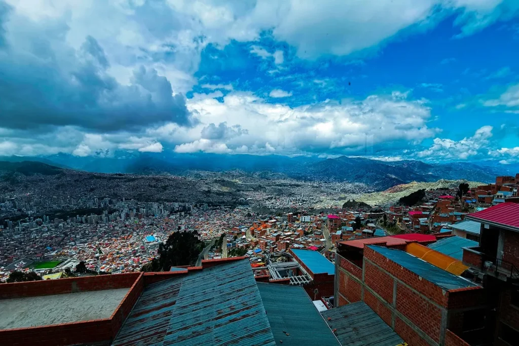 Vista ampla de La Paz, na Bolívia, observada do alto de uma colina, com casas de tijolos no primeiro plano e a cidade se estendendo até as montanhas sob um céu azul com nuvens densas.