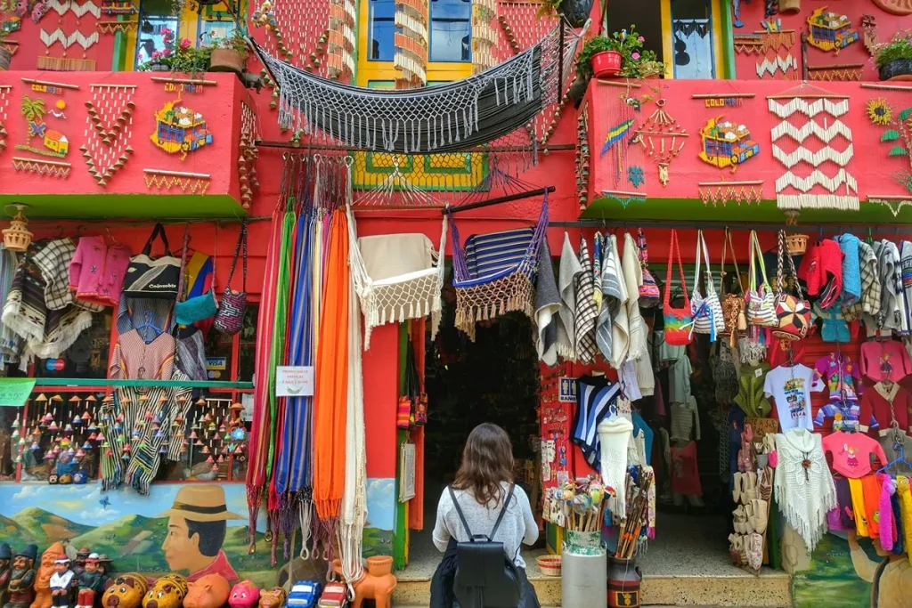 Fachada de uma loja de artesanato na Colômbia, com roupas, bolsas, redes e objetos coloridos expostos, e uma visitante observando a entrada.