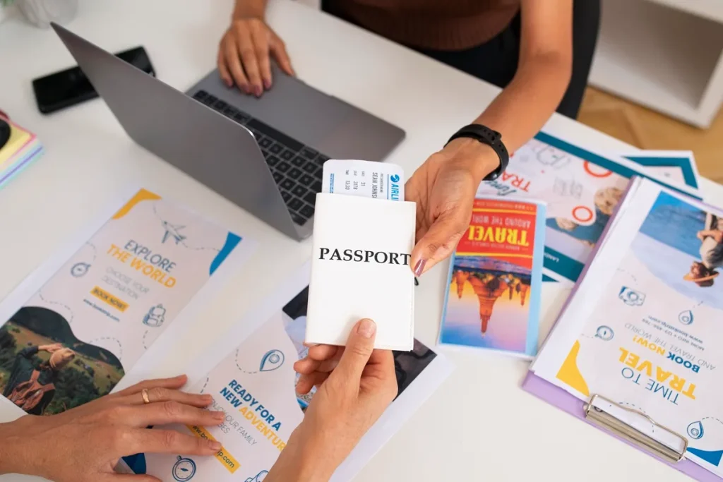 Pessoa entregando passaporte com passagem aérea a uma agente de viagens em mesa com panfletos de turismo e notebook.