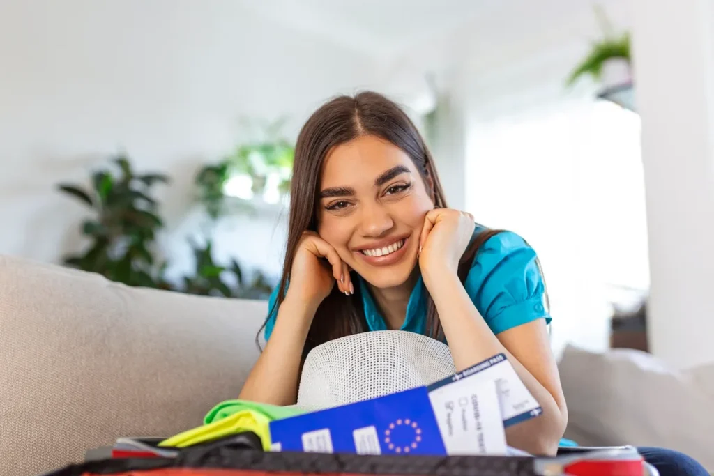 Mulher sorrindo ao lado de mala aberta com passaporte, passagens e acessórios de viagem, em ambiente iluminado.