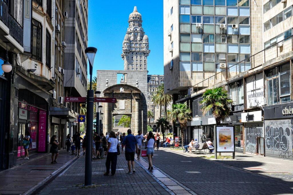 Pedestres caminhando por rua histórica de Montevidéu com edifícios comerciais e monumentos ao fundo.