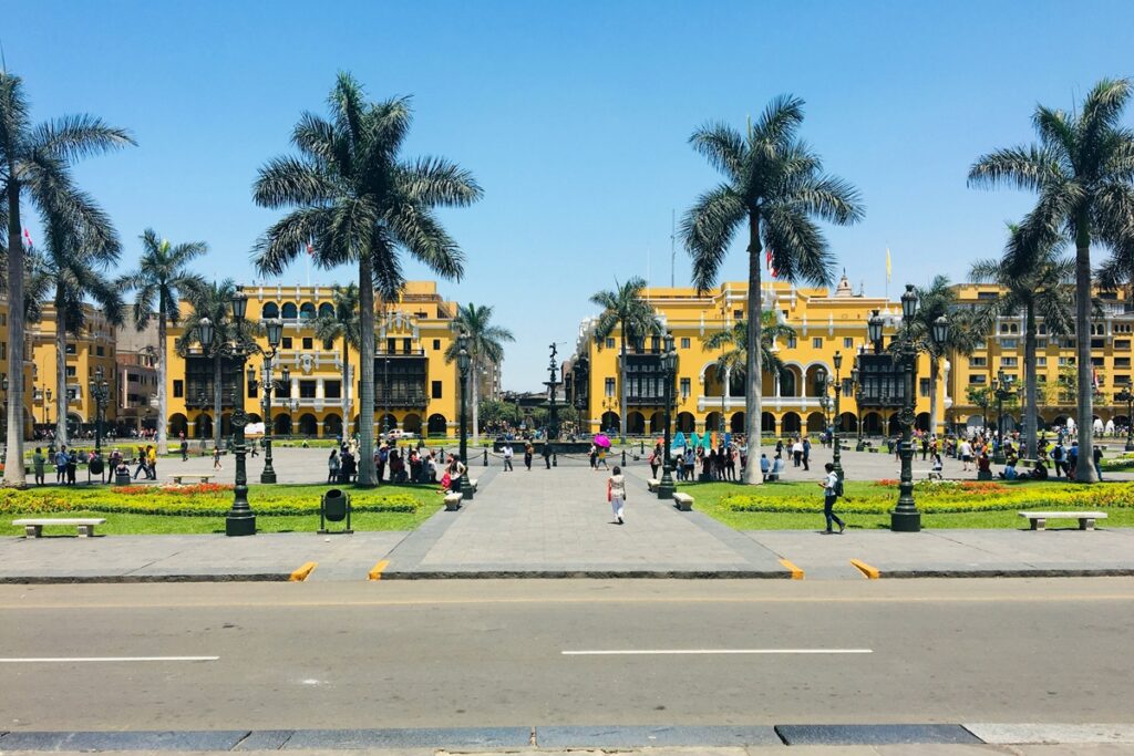 Vista da Plaza Mayor de Lima com edifícios coloniais amarelos, palmeiras e visitantes caminhando pelo centro histórico da capital peruana.